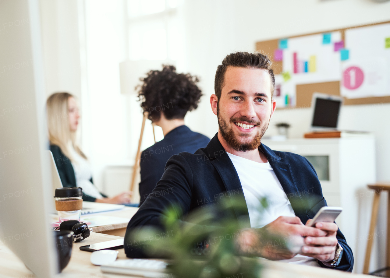 A portrait of young hipster businessman with smartphone and colleagues in the background in a modern office.