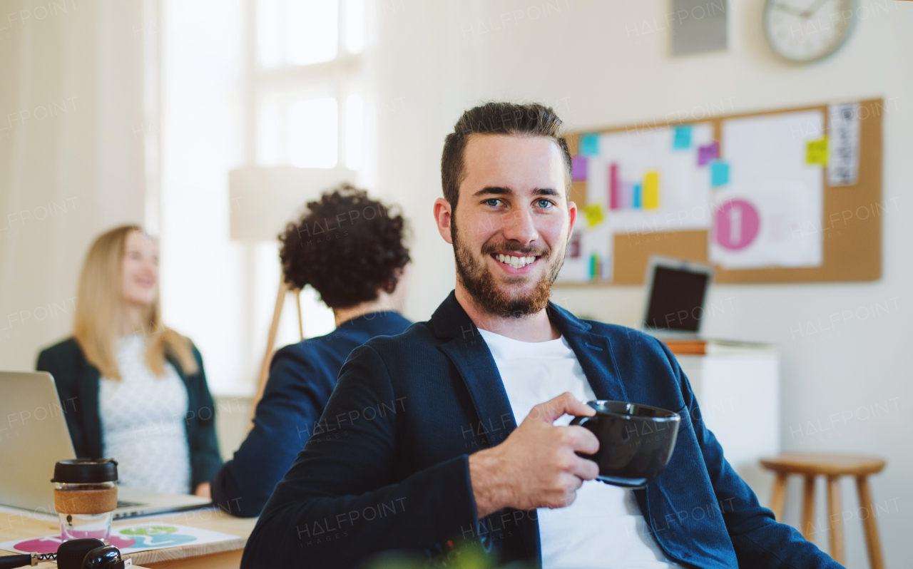 A portrait of young hipster businessman with coffee and colleagues in the background in a modern office.