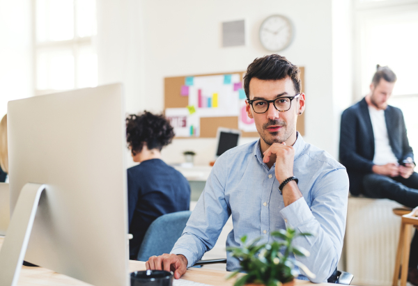 A portrait of young businessman with desktop computer working in a modern office, colleagues in the background.