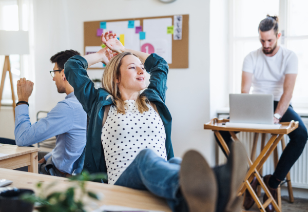 Portrait of young relaxed businesswoman with colleagues in a modern office, hands behind head and feet on desk.