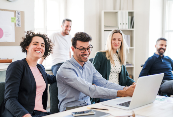 Group of young, cheerful businesspeople with laptop sitting and standing around table in a modern office, having meeting.