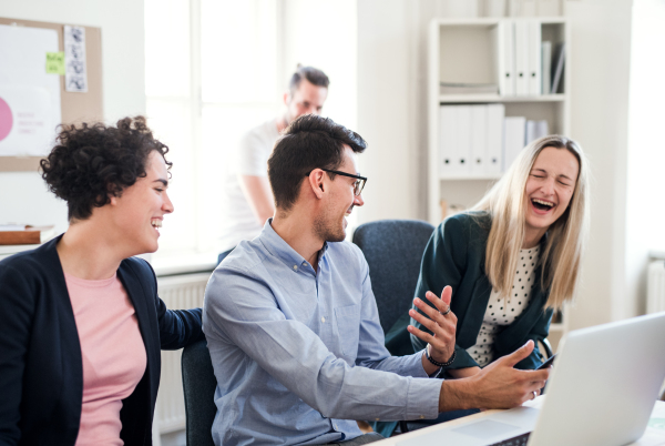 Group of young, cheerful businesspeople with laptop sitting around table in a modern office, laughing when having meeting.