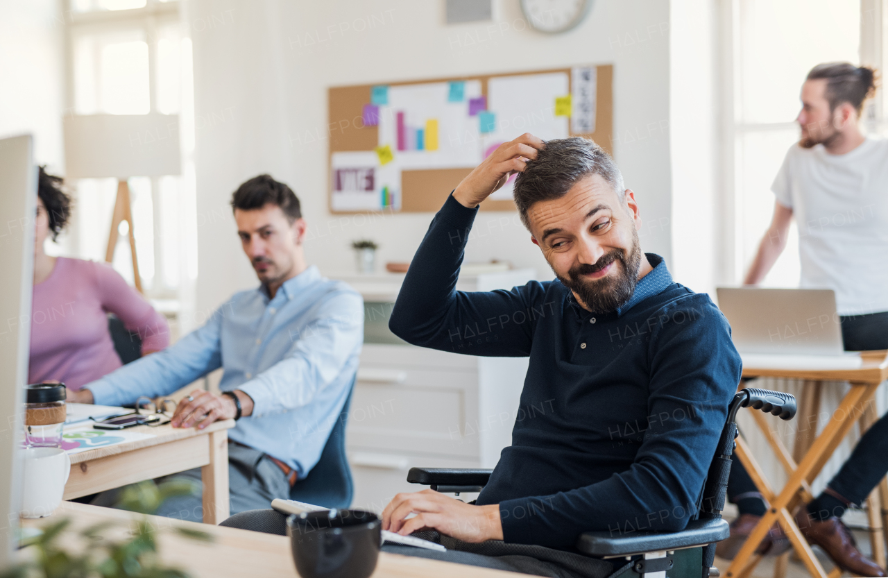 Young businessman in wheelchair with colleagues in a modern office, scratching head.