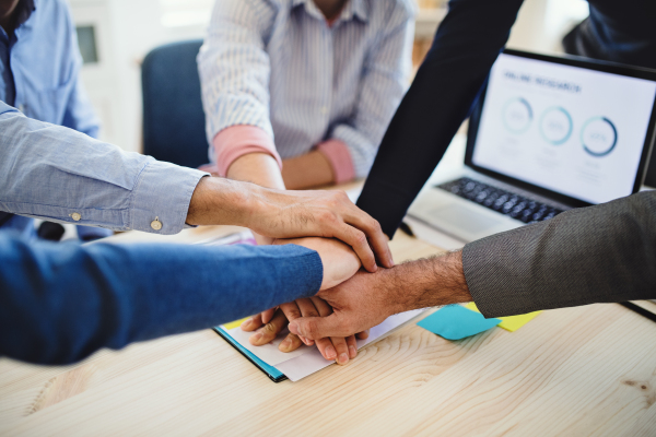 A midsection of young businesspeople sitting around table in a modern office, putting hands together. A hand-stack gesture.