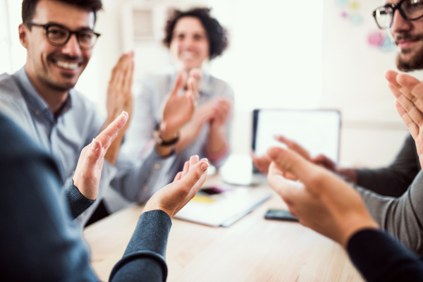 Group of young, cheerful businesspeople sitting around table in a modern office, clapping.