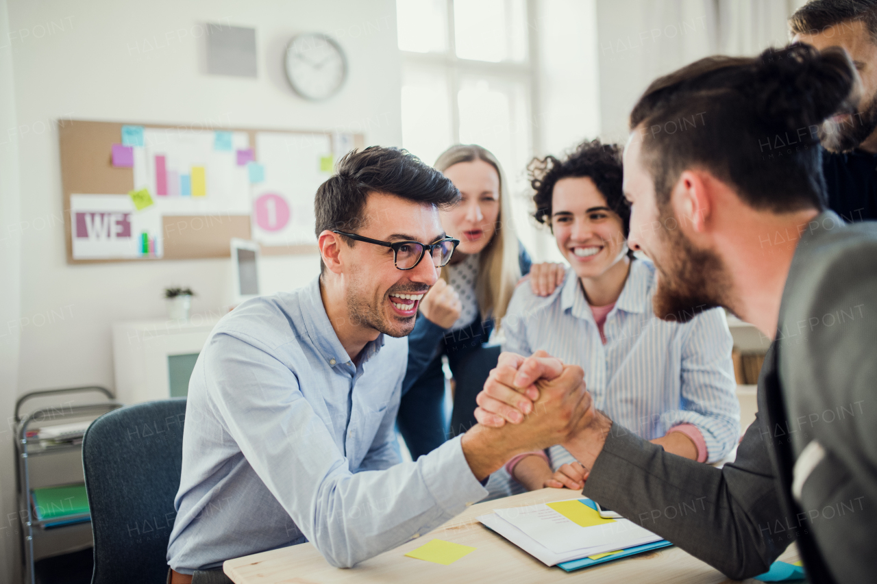 Two businesspeople with colleagues in the background in modern office, shaking hands.