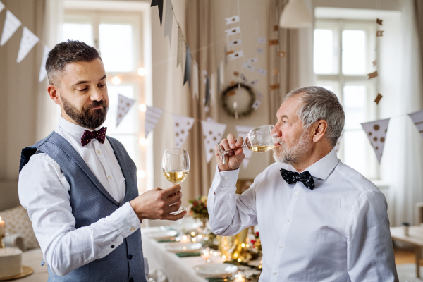 A portrait of a senior and hipster mature man standing indoors in a room set for a party, degusting wine.