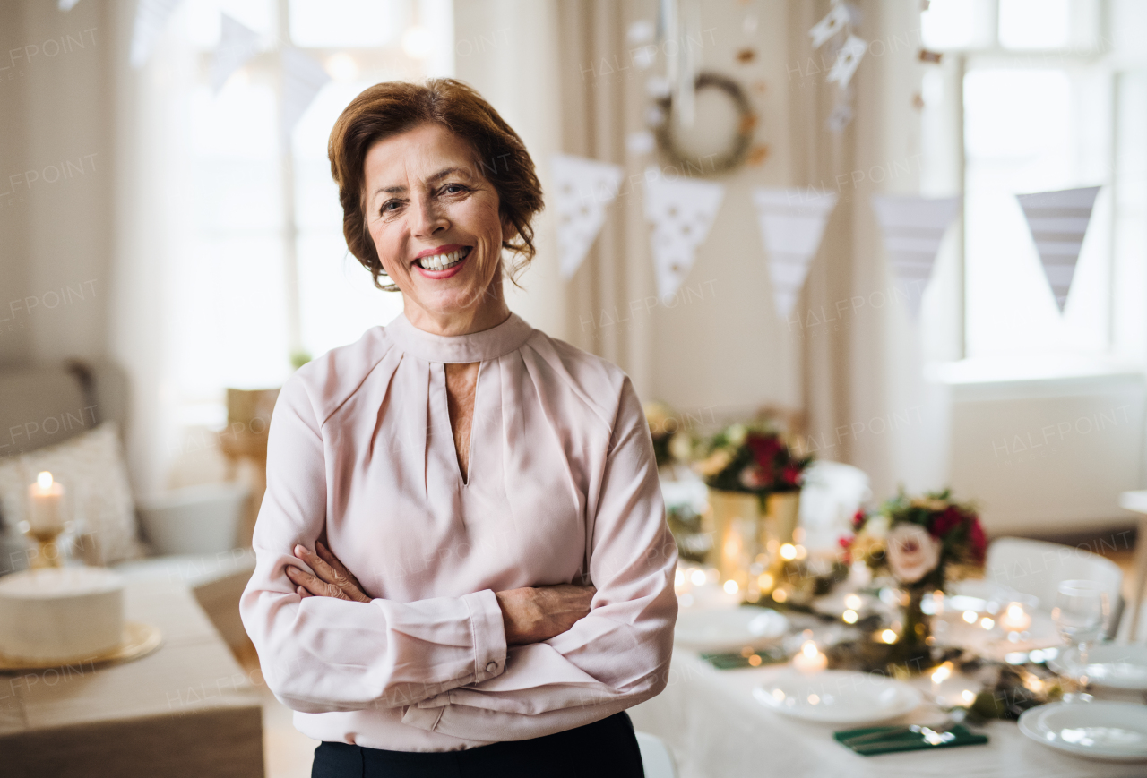 A portrait of a senior woman standing indoors in a room set for a party, arms crossed.