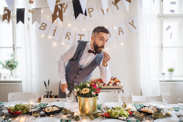 A mature man standing indoors in a room set for a party, making funny faces and nibbling at food.