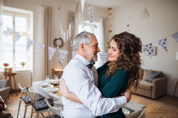 A portrait of a senior man and young woman standing indoors in a room set for a party, hugging.