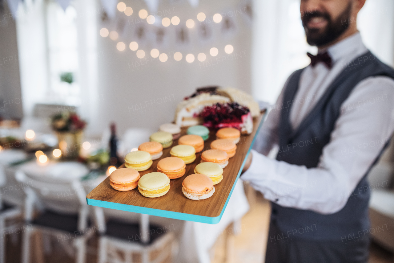 A midsection of man standing indoors in a room set for a party, holding a tray with biscuits.