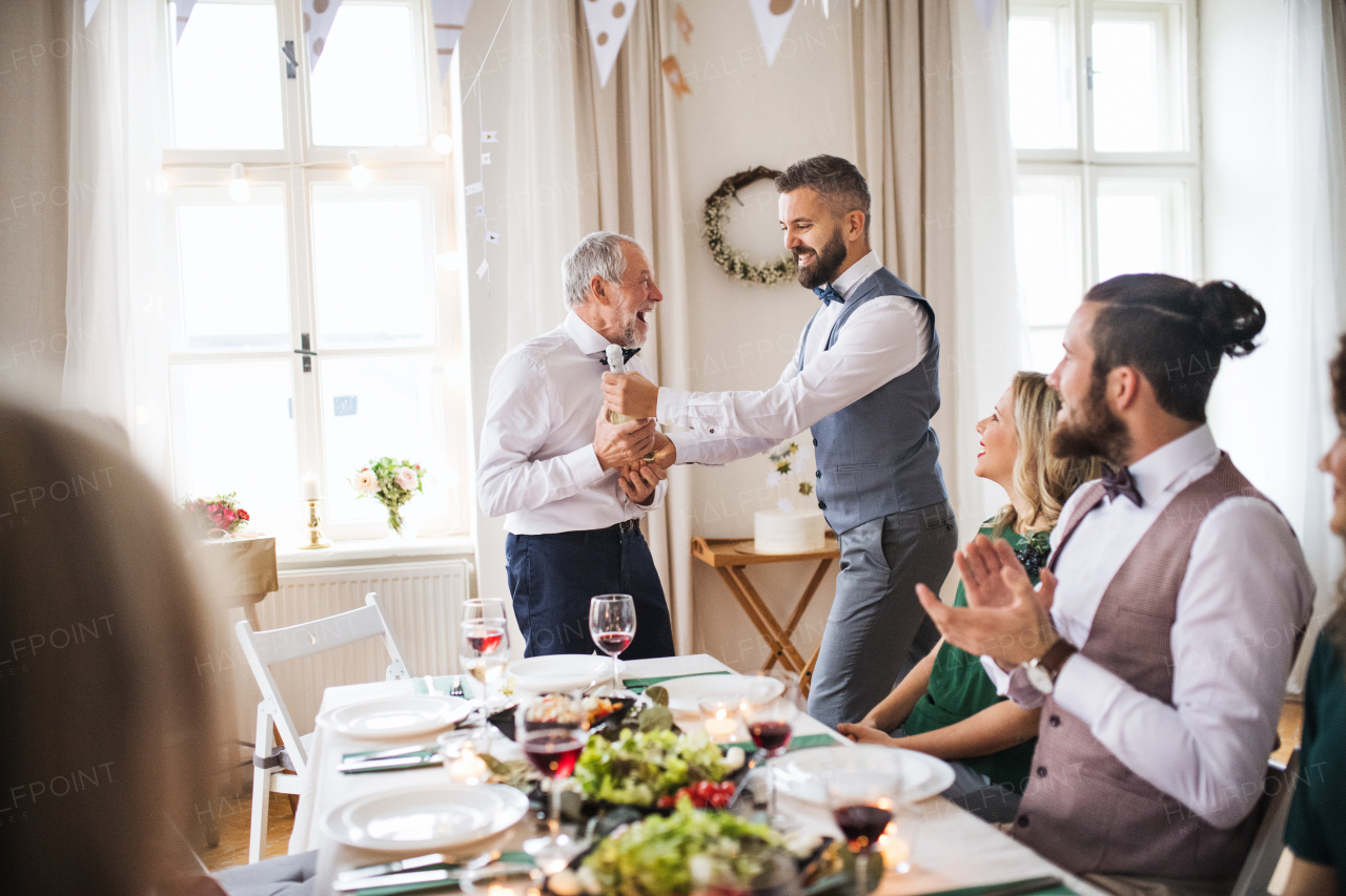 A mature man giving a bottle of wine to his father on indoor birthday party, a celebration concept.