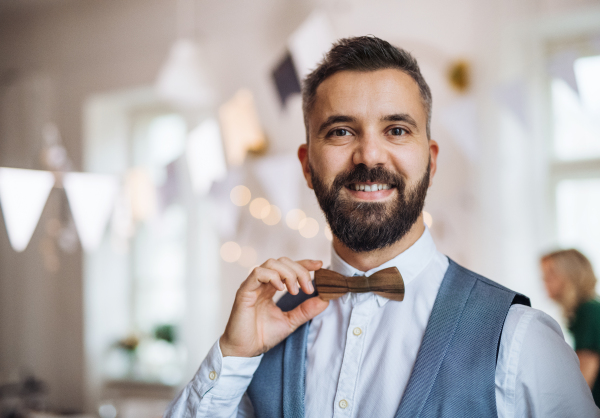 A portrait of a hipster mature man standing indoors in a room set for a party, holding wooden bowtie.