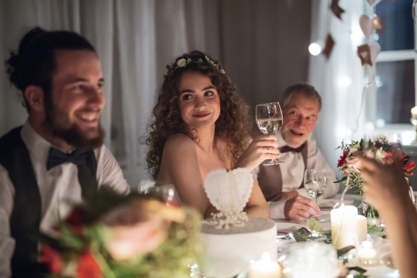 A young happy couple sitting at a table on a wedding, talking to guests.