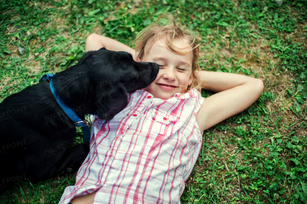 A small girl with closed eyes lying down on grass, a black dog licking her face.