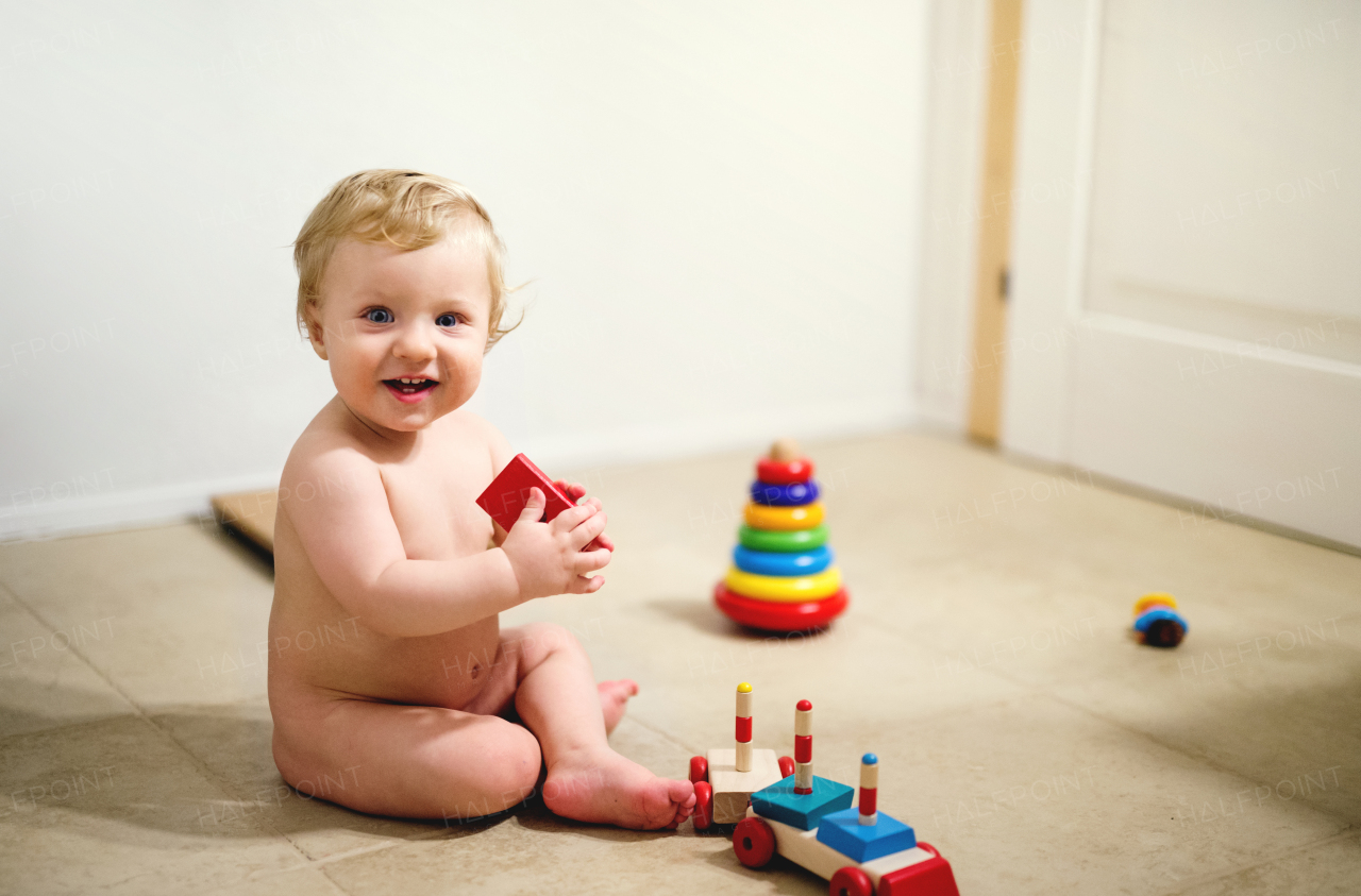 A happy naked toddler boy sitting on the floor at home, playing with colored wooden trian toys.