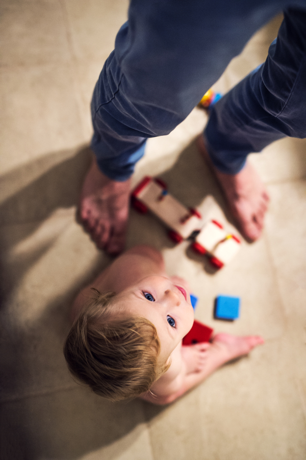 A naked toddler boy with his unrecognizable father sitting on the floor at home, playing with wooden blocks. Top view.