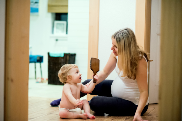 Beautiful pregnant woman sitting with a baby boy on the floor at home. A toddler child holding a comb.