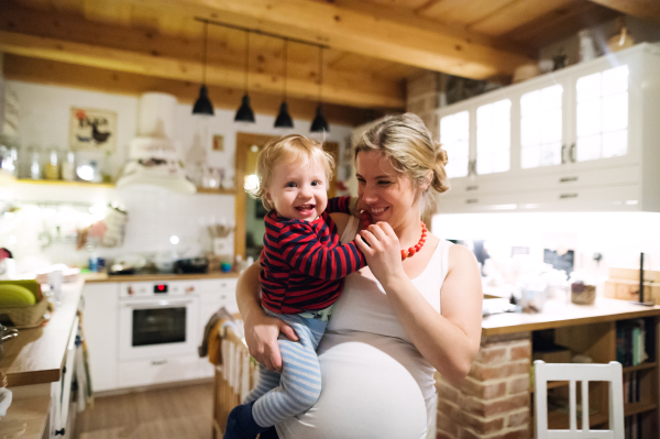 Beautiful young pregnant woman carrying a toddler boy in the kitchen at home.