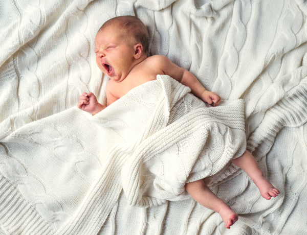 Cute newborn baby lying and yawning on bed, covered by a white blanket. Top view.