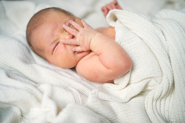 A close-up of a newborn baby with a hand on her or his face lying on bed, covered by a white blanket.