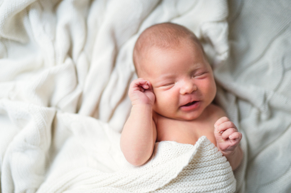 A close-up of a cute newborn baby lying on bed, covered by a white blanket, sleeping and smiling. Top view.