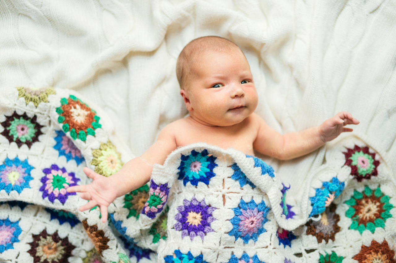 A close-up of a cute happy newborn baby lying on bed, covered by a colorful crocheted blanket. Top view.