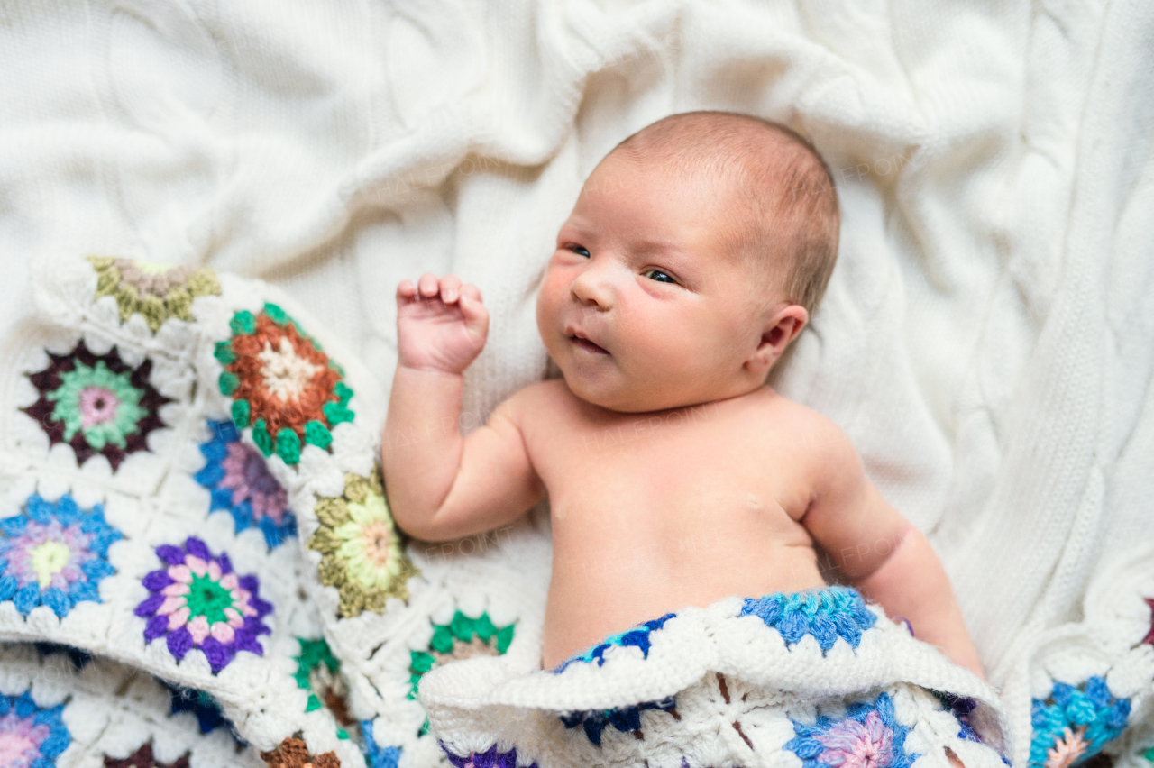 A close-up of a cute happy newborn baby lying on bed, covered by a colorful crocheted blanket. Top view.