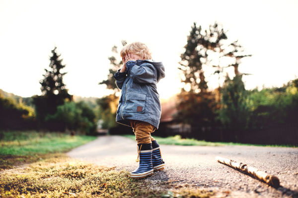 A little toddler boy standing outdoors on a road at sunset, covering his eyes. Copy space.