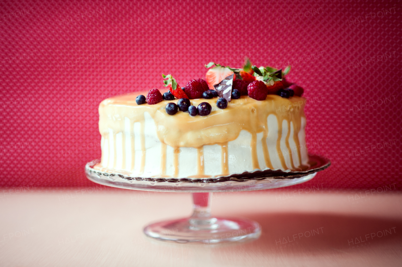 A birthday cake on a glass stand on the table, fruit on top. Red background.