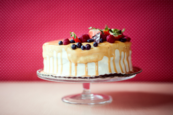 A birthday cake on a glass stand on the table, fruit on top. Red background.