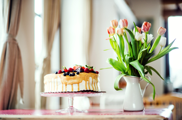 A composition of a birthday cake on a glass stand and a vase with tulips on the table.