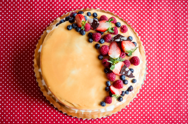 A birthday cake on a glass stand on the table, fruit on top. Red background. Top view.