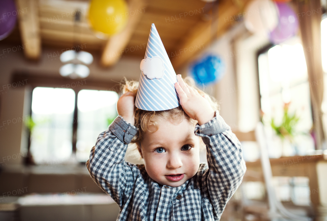 A happy toddler boy with a party hat standing inside at home.