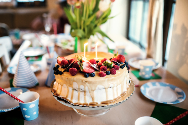 A birthday cake on a glass stand and a vase with tulips on the table set for a birthday party.