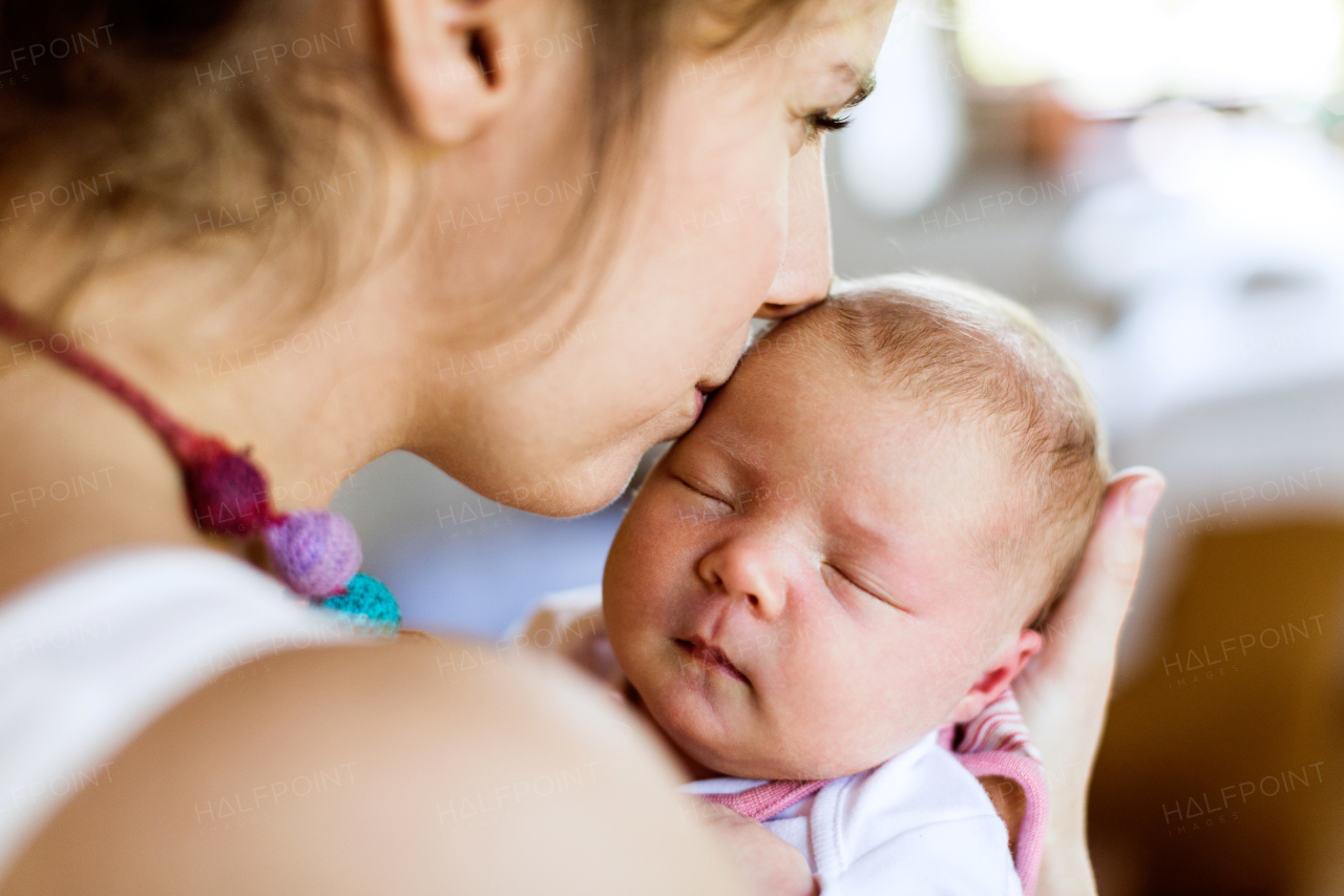 Beautiful young mother at home holding her cute newborn baby girl, kissing her on forehead.