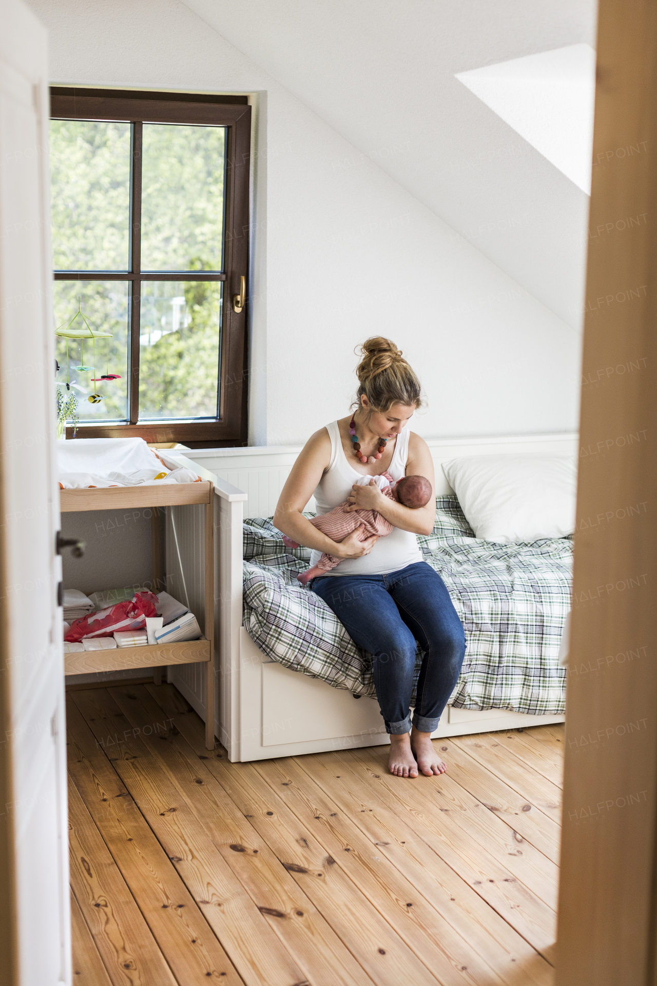 Beautiful young mother at home sitting on bed holding her cute newborn baby girl, kissing her on head.