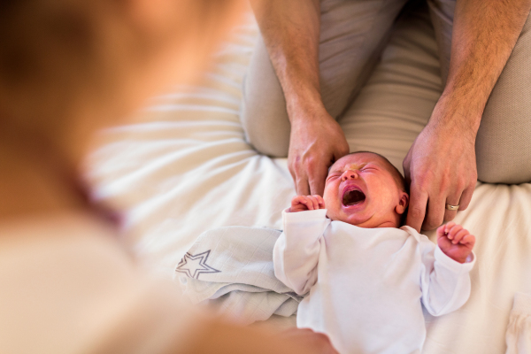 Unrecognizable young parents at home dressing their cute baby daughter on bed, little girl crying.