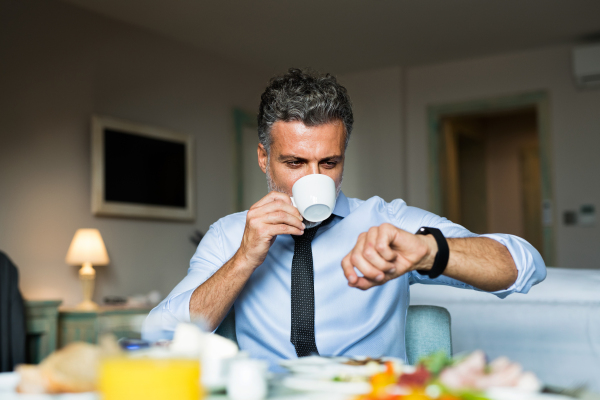 Mature, handsome businessman having breakfast in a hotel room, drinking cofee and checking time.