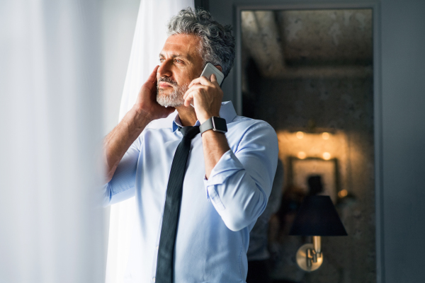 Mature businessman with smartphone in a hotel room. Handsome man standing at the window, making a phone call. Close up.