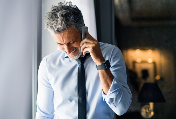 Mature businessman with smartphone in a hotel room. Handsome man standing at the window, making a phone call. Close up.