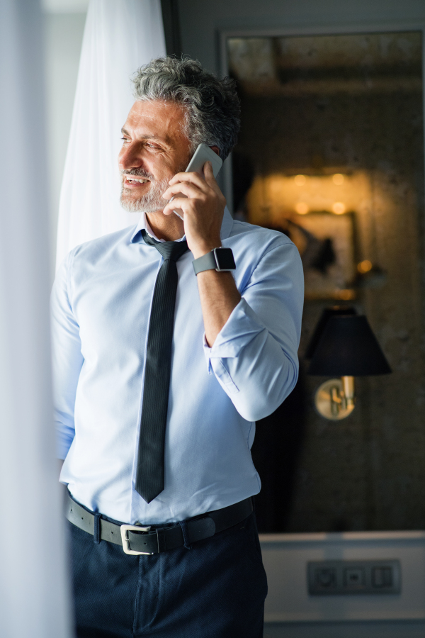 Mature businessman with smartphone in a hotel room. Handsome man standing at the window, making a phone call.