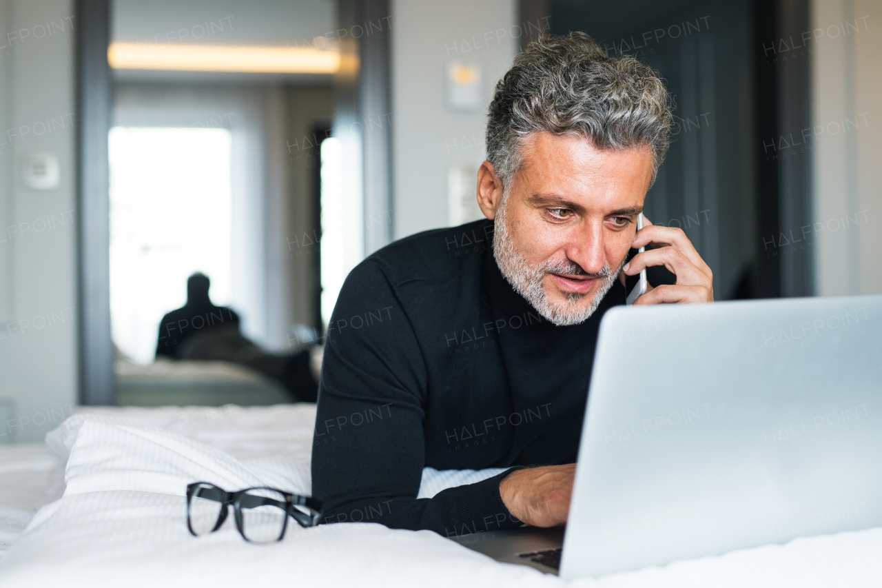 Mature businessman with laptop and smartphone in a hotel room. Handsome man working on computer and making a phone call.