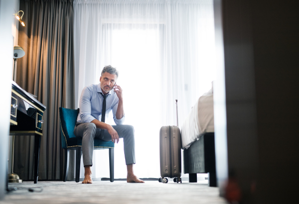 Mature businessman with smartphone in a hotel room. Handsome man sitting on a chair, making a phone call.