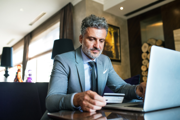 Handsome mature businessman with laptop in a hotel lounge. Man holding a credit card. Internet banking. Online payment.