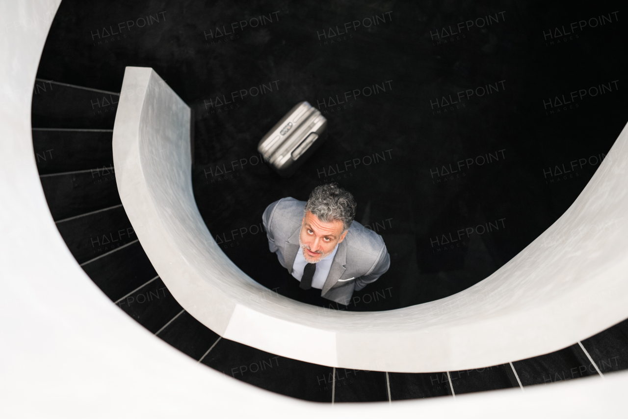 Businessman with suitcase standing at the bottom of the staircase, looking up. Top view.