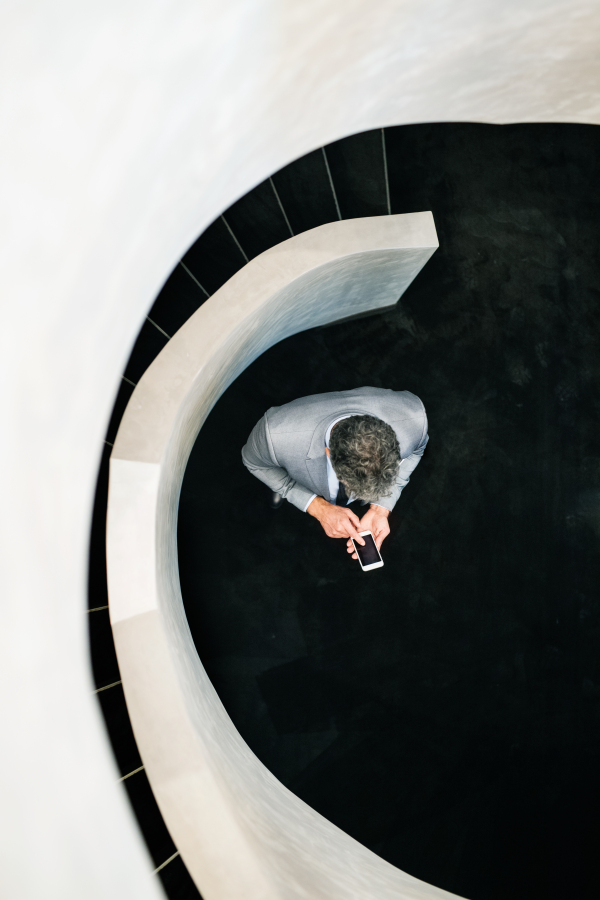 Businessman with smartphone and suitcase standing at the bottom of the staircase, texting. Top view.