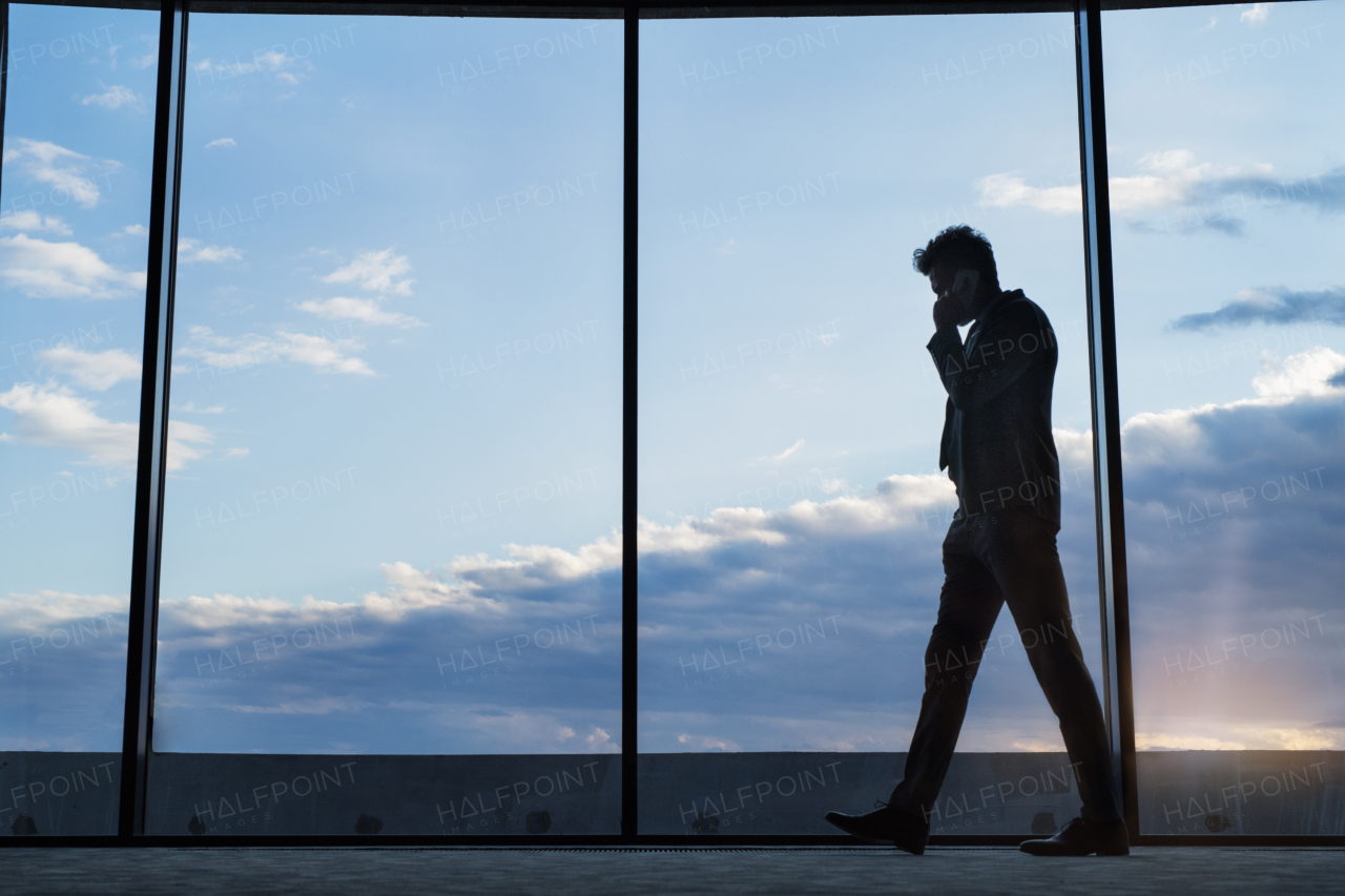 Mature businessman in a hotel with a smartphone. Man making a phone call, walking along big windows.