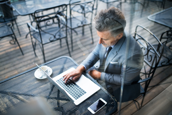 Mature businessman with laptop and smartphone sitting outside a cafe. Man working on a laptop. Shot through glass.