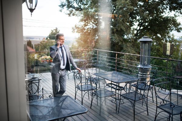 Mature businessman with smartphone standing in an outdoor hotel cafe. Man making a phone call. Shot through glass.